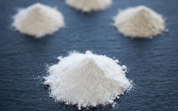 A single scoop of creatine monohydrate in sharp focus in the foreground, with three smaller scoops of alternative creatine forms blurred in the background on dark slate