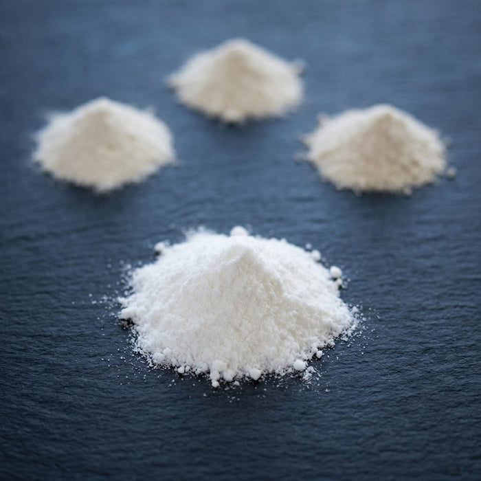 A single scoop of creatine monohydrate in sharp focus in the foreground, with three smaller scoops of alternative creatine forms blurred in the background on dark slate