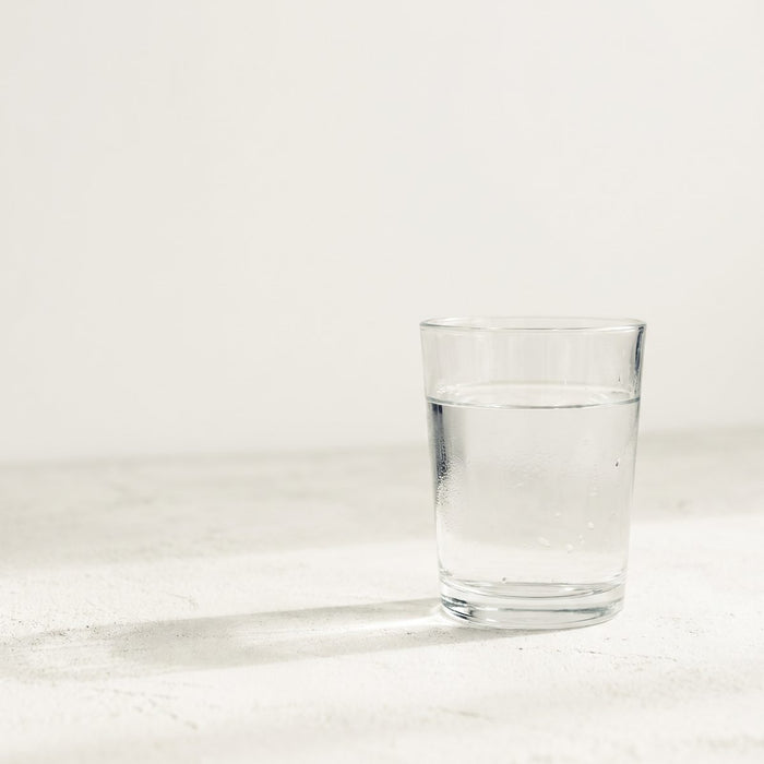 A single pint glass of clear water on a pale concrete surface in soft morning light, condensation on the glass, nothing else in frame